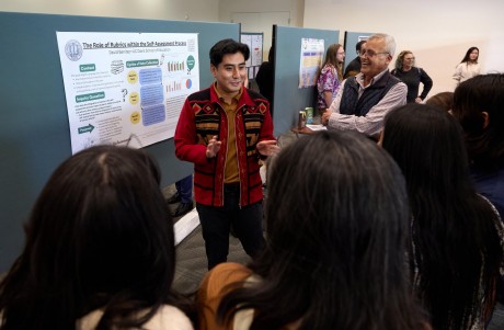 A student in a red patterned jacket and a brown shirt stands in front of a research poster talking to a group of people.