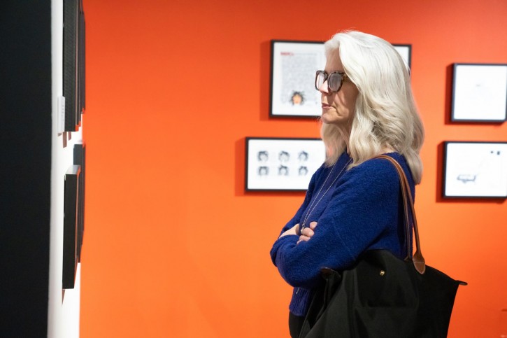 A person with a vibrant blue sweater, a black bag, and light blonde hair admires the art on the wall.