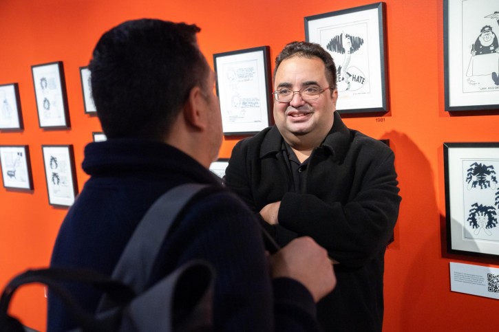 Enrique García stands in front of art on an orange wall talking to another person.
