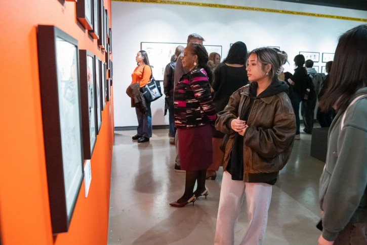 A collection of people stand in front of frames of art on an orange wall.