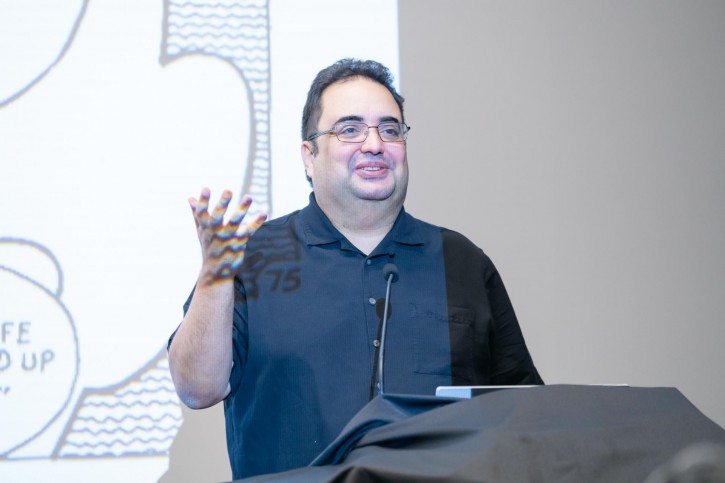 Enrique García stands in front of a podium and a projector screen addressing the audience.