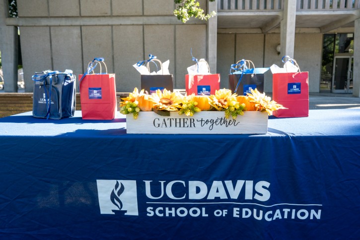 Blue, red, and brown gift bags are lined up on a table that has fall decor and a School of Education dark blue tablecloth.