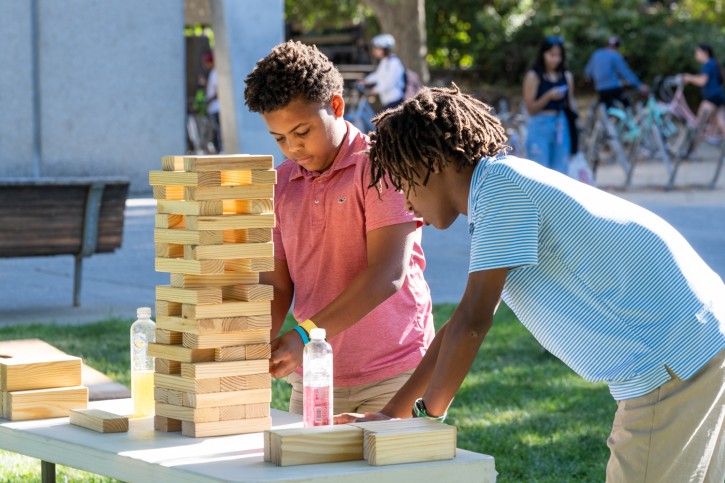 A child wearing a blue shirt studies the tower of Jenga blocks while a child in a red shirt pulls a block out from the center of the tower.