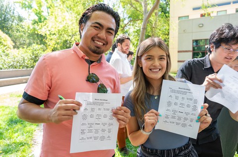 Two people pose outdoors for the camera while holding up pieces of paper.