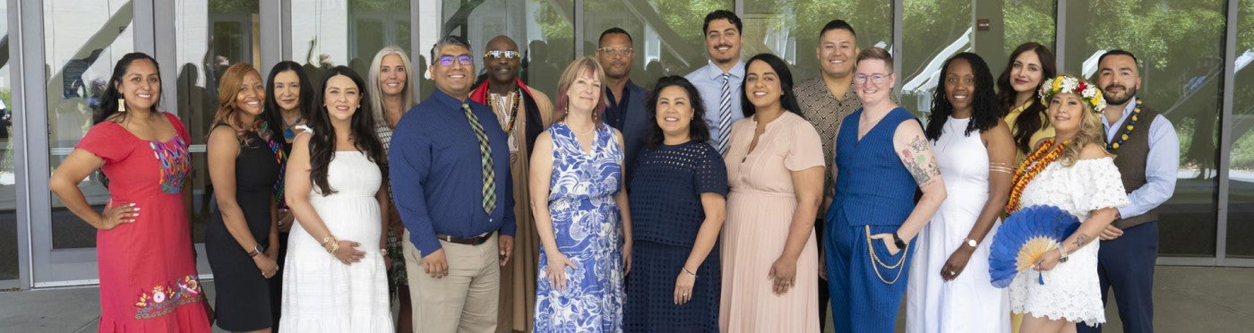 Group portrait of the 2026 CANDEL graduating cohort standing outside a campus building. Eighteen graduates pose together in two rows in front of the Conference Center, smiling at the camera. They wear a mix of colorful dresses, dress shirts, ties, leis, and cultural regalia, reflecting the diversity of the cohort.