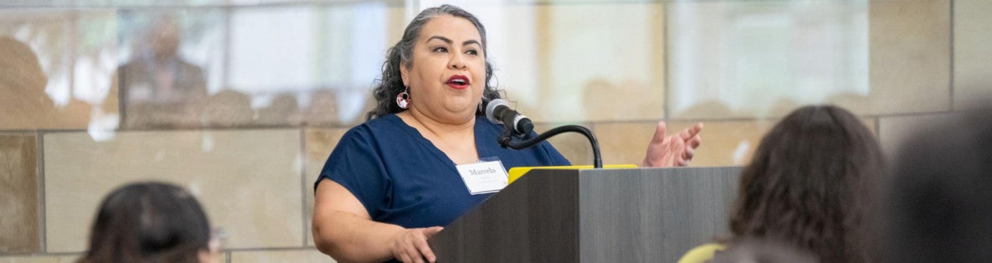 Marcela Cuellar stands at a podium and addresses an audience seated in front of her.