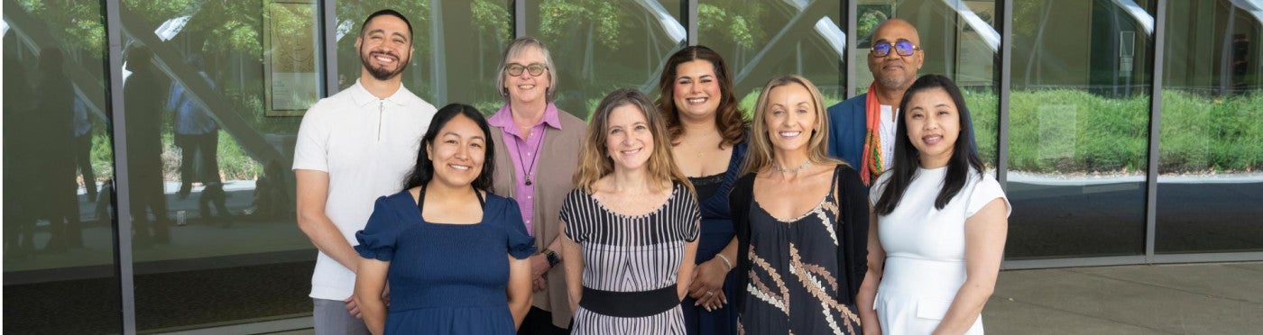 Eight PhD graduates standing in two rows in formal clothing on graduation day