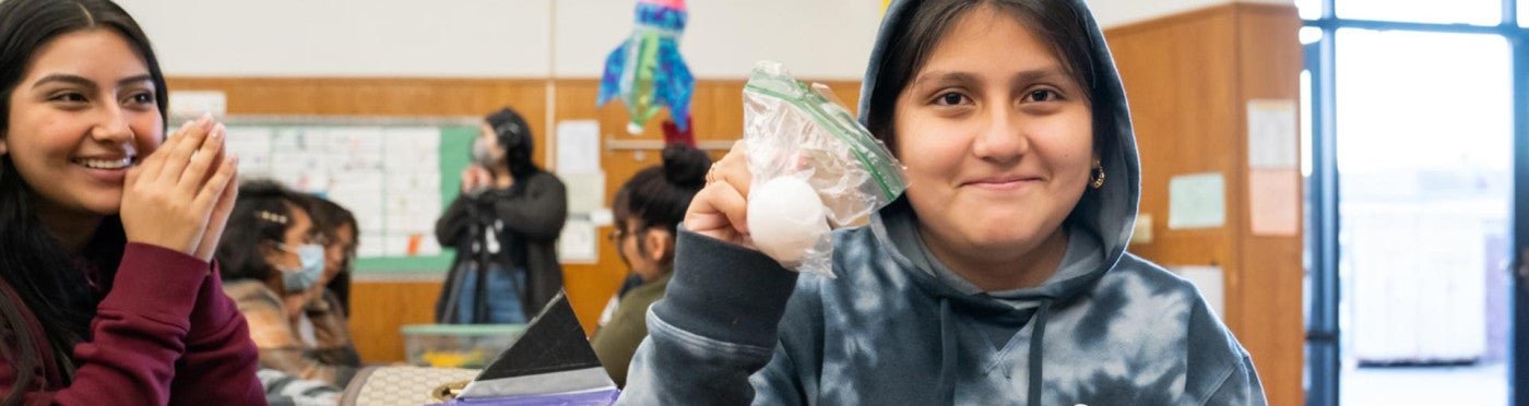 Satisfied looking student in a hoodie holds up a science project as another student looks on and smiles
