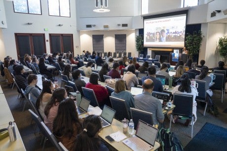 A large group of people sit facing a presentation screen.