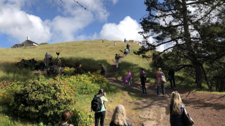 A line of people wearing backpacks walk up a grassy hill with blue skies and clouds in the background.