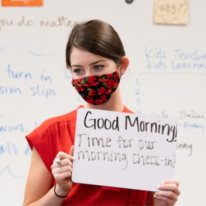 An elementary school teacher wearing a mask holds a sign saying Good Morning