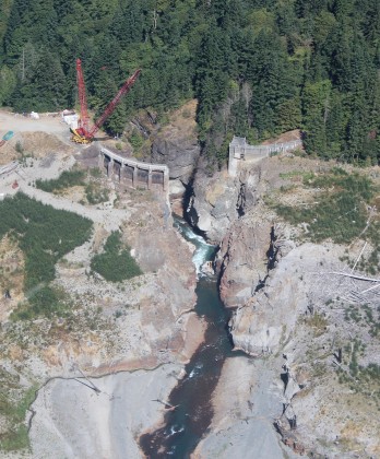 Aerial photograph of the Elwha River flowing through the remains of the Glines Canyon Dam during the 3rd year of the dam removal project.  A crane sits to the left of the remains of the dam, and the river flows through a  steep canyon past it.