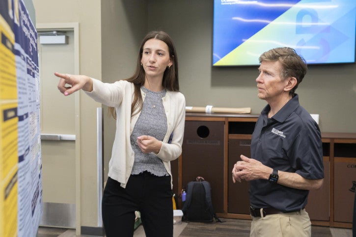 A young woman gestures at a poster while explaining her research to Dean Tom Smith.