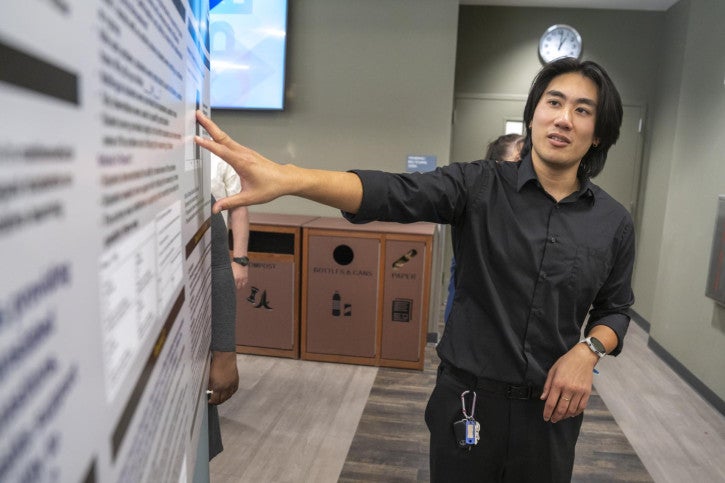 A man in a black shirt is presenting a large research poster, gesturing with one hand.