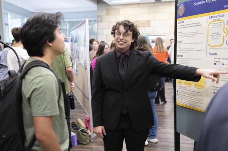 A person in a suit happily presents a research poster to an interested onlooker. 