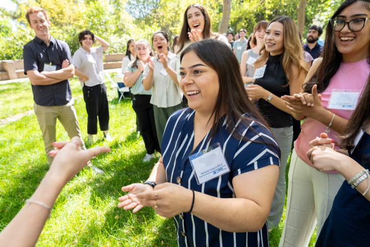 A student is playing rock paper scissors with another student out of frame. Their classmates cheer around them.