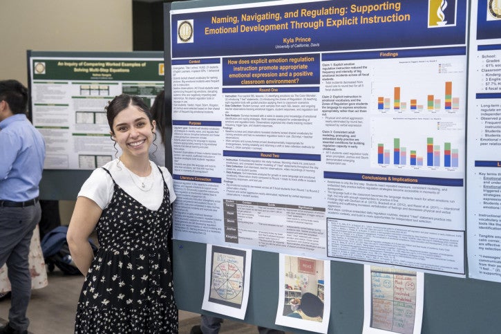 A woman in a black floral dress stands smiling beside a large academic poster on emotional development in education, with colorful charts and text.