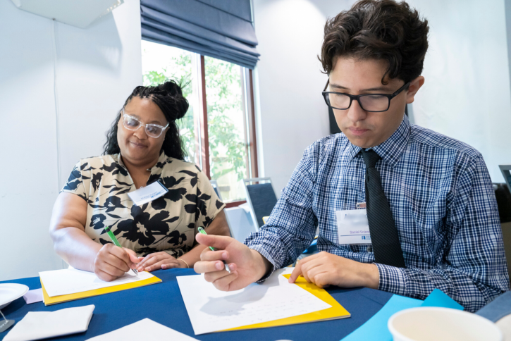 Two students sit at a table, writing.