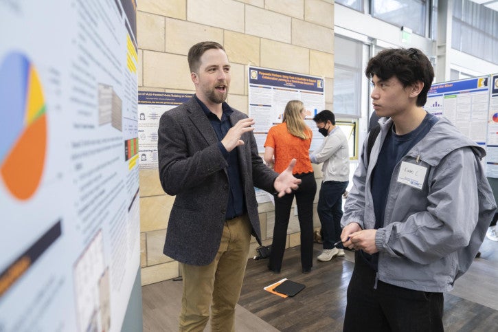 A man passionately explains his research. A young man listens intently.