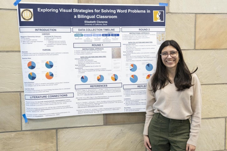 A woman with long dark hair and glasses stands smiling in front of a research poster titled "Exploring Visual Strategies for Solving Word Problems in a Bilingual Classroom." The poster features charts and text sections, highlighting data collection and analysis. 
