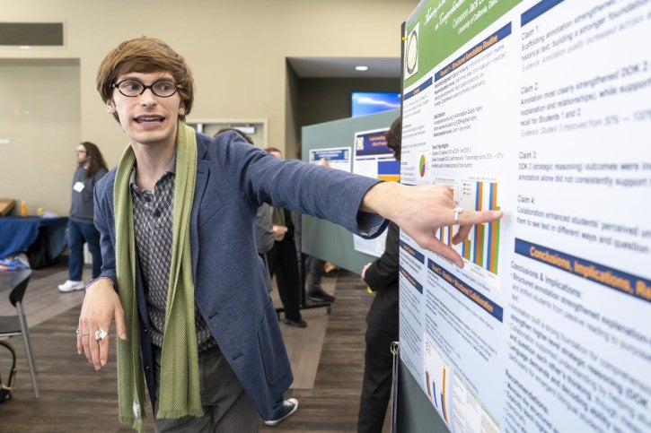 Person with glasses and a green scarf excitedly presents research on a poster board. They point to colorful graphs.