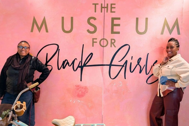 Two women lean against a pink wall that reads "The Museum for Black Girls"