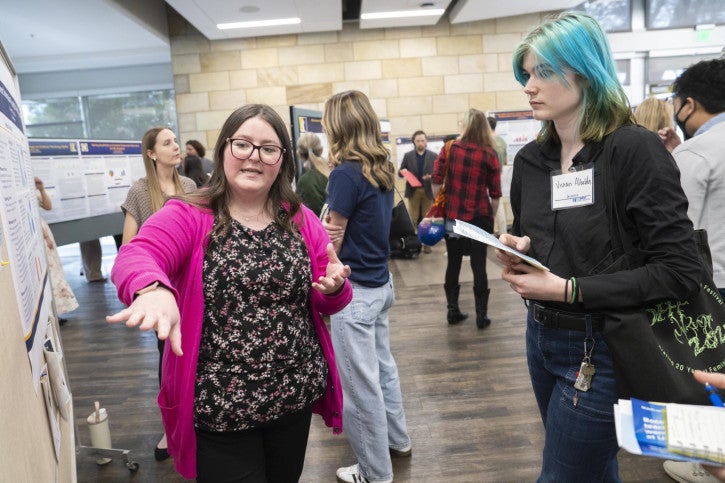 A woman in a pink cardigan passionately presents research on a poster. Attendees listen, take notes, and engage in discussion.