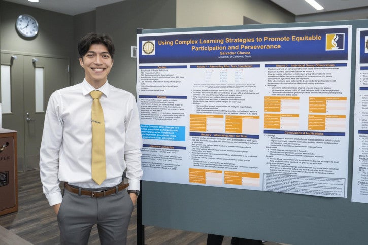 A smiling person in a tie stands beside an academic poster titled "Using Complex Learning Strategies to Promote Equitable Participation and Perseverance."