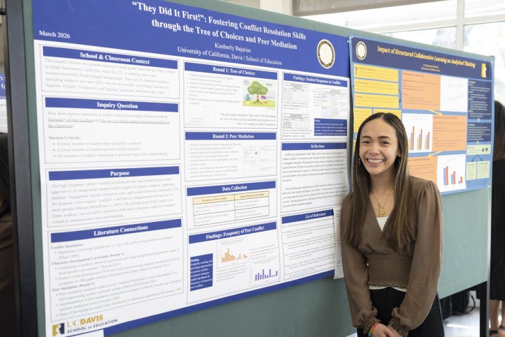 Smiling woman stands beside a detailed academic poster on conflict resolution, showcasing graphs and text