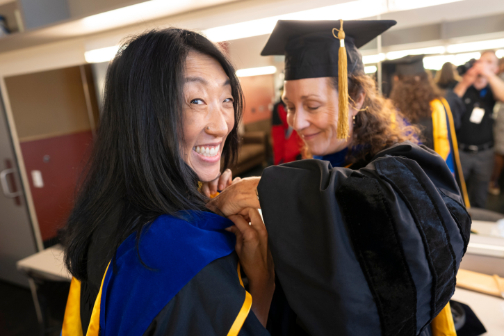Lisa Sullivan helps Nancy Tseng with her regalia while Nancy smiles at the camera.