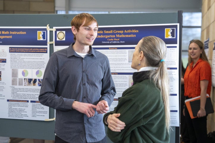 A young man talks with an older woman in front of academic posters at a presentation. A woman in orange stands in the background, holding a notebook.