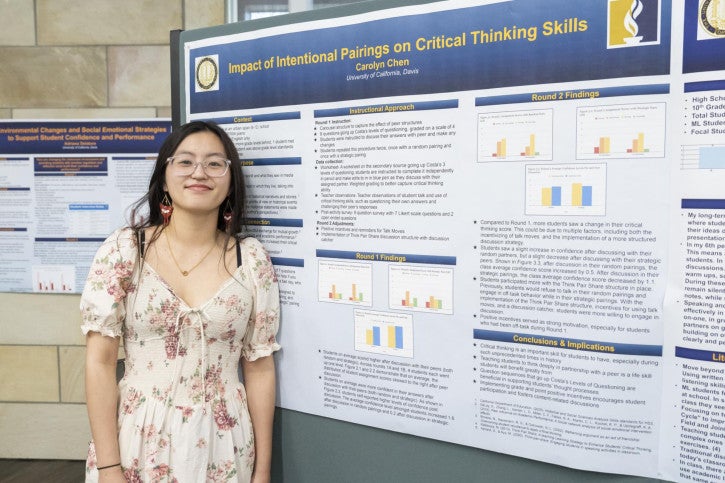 A woman in a floral dress smiles next to a research poster titled "Impact of Intentional Pairings on Critical Thinking Skills," featuring charts and detailed text.