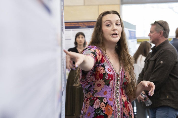 Woman in a floral dress points at a poster in a crowded room, explaining her research. 