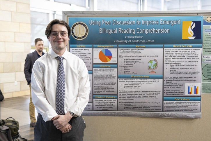 Man in a white shirt and tie stands smiling in front of a research poster titled "Using Peer Discussion to Improve Emergent Bilingual Reading Comprehension."
