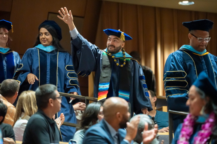 A PhD grad waves to someone in the audience as he walks into Jackson Hall.