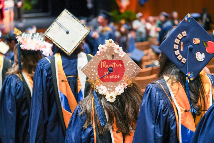 MA graduates walk to their seats, sporting highly decorated grad caps.
