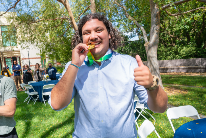 A student with brown curly hair and a mustache pretends to bite a fake gold medal.