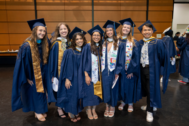 A group of MA graduates wearing navy blue regalia smile for the camera. Some wear gold stoles or colorful stoles decorated by their students.