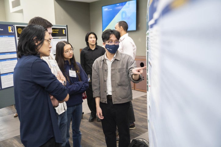 A man wearing a face mask presents a poster to a group listening attentively.