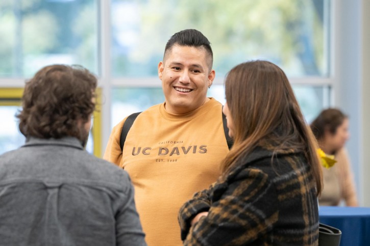 A man wearing a yellow UC Davis sweatshirt smiles to two women who are facing away from the camera.