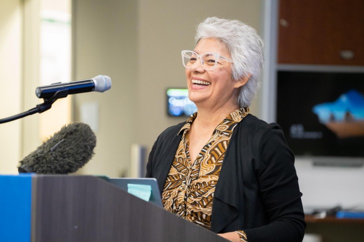 Gloria Rodriguez stands at the podium, faces the audience, and speaks into a microphone.