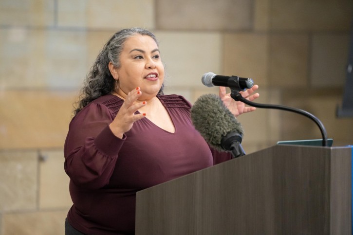 Marcela Cuellar stands at the podium, faces the audience, and speaks into a microphone.