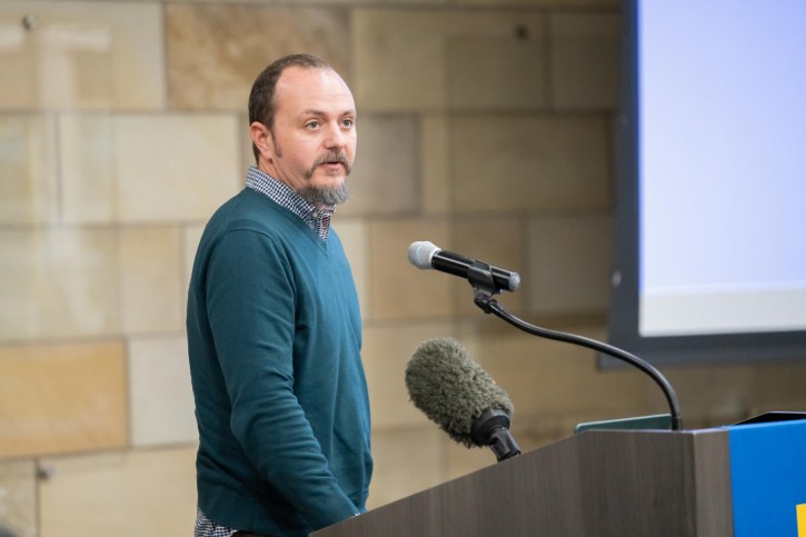 Jacob Hibel stands at the podium, faces the audience, and speaks into a microphone.