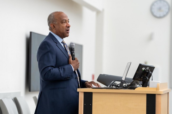 Gary May stands at a podium, addressing an audience sitting in front of him.