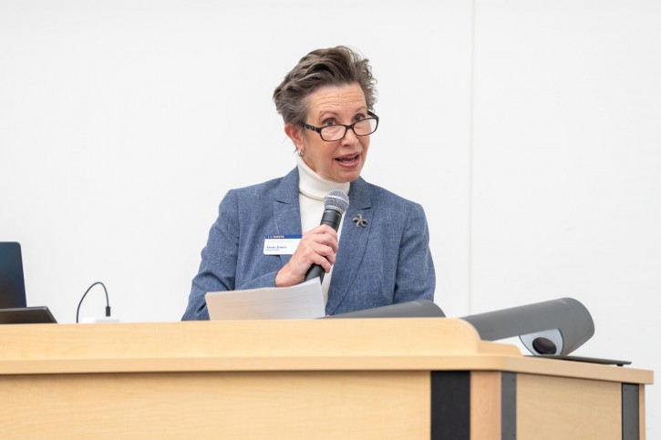 Anne Jones stands at a podium, addressing an audience sitting in front of him.