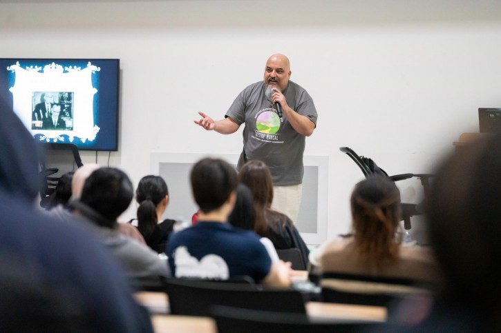 Samuel Blanco III stands on stage, addressing an audience sitting in front of him.