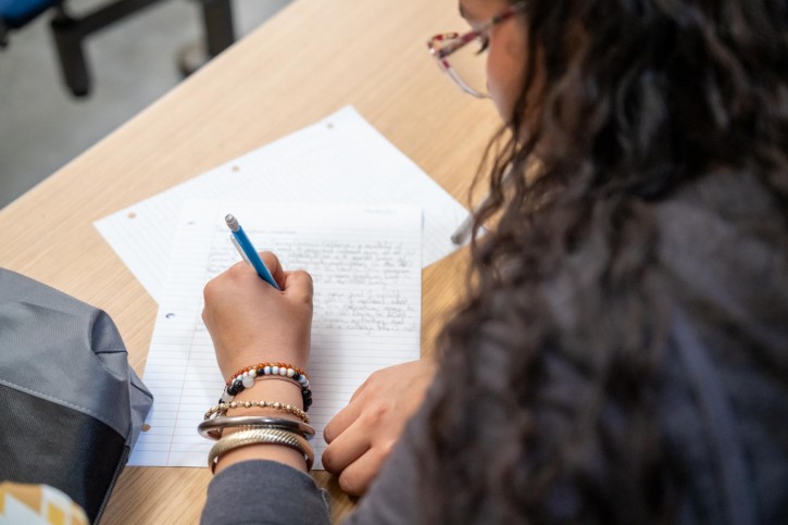 A girl holds a pen in her left hand and writes on lined paper.