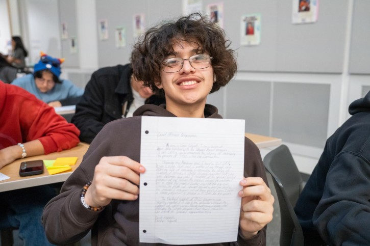 A boy holds up a piece of lined paper where he's written a letter on it.