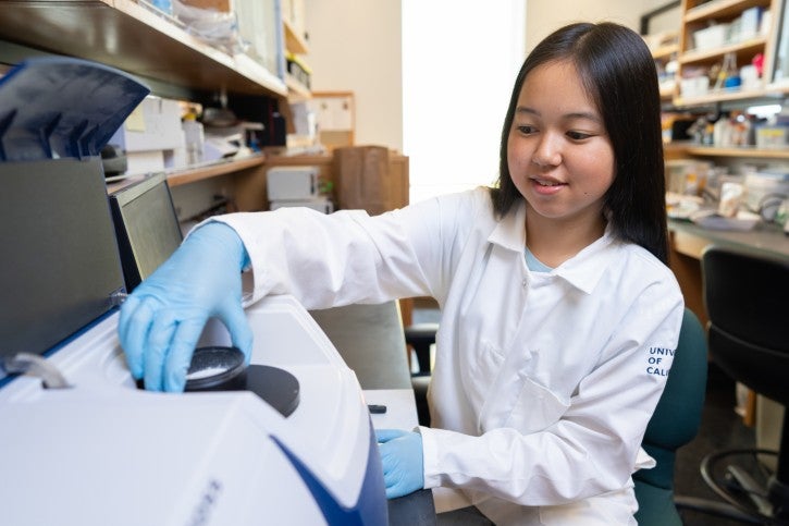 Student wearing gloves and white lab coat puts sample into machine in lab
