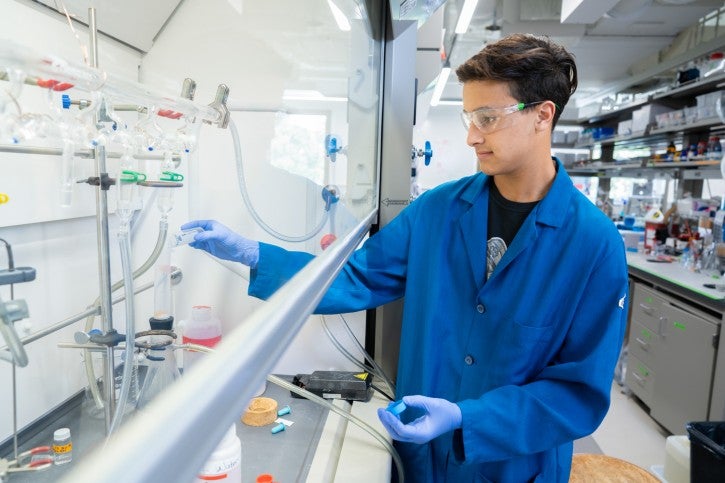 Student wearing gloves and blue lab coat works under lab hood